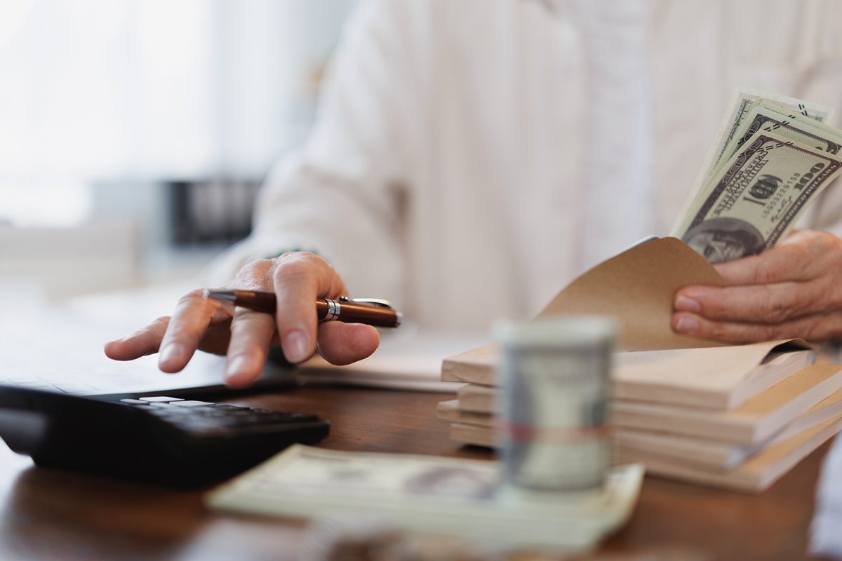 Senior accountant calculating and counting American one hundred dollar bills at an office desk, utilizing a calculator, pen, and notebook for precise financial management and record-keeping